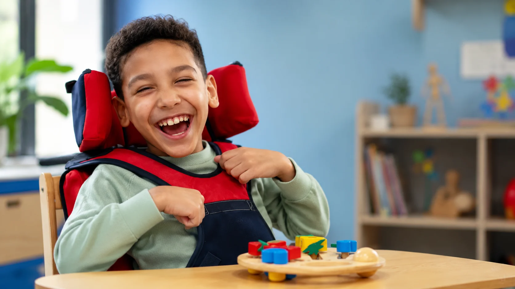Menino sorrindo em cadeira adaptada, brincando com um encaixe de madeira colorido sobre a mesa da sala de aula.